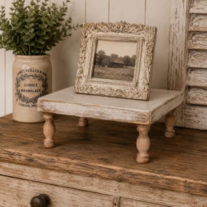 May include: A square wooden riser with a distressed white finish and four turned light brown legs. Resting on the riser is an ornate, rectangular picture frame with a cream-colored finish containing a black and white photograph of a barn.