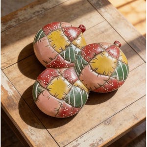 May include: Three colorful, round decorative objects with a patchwork design. Each object features a combination of red, yellow, green, and pink patches, with white dotted accents. They are arranged on a light-colored wooden surface.