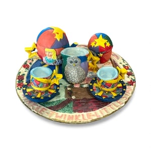 May include: A whimsical ceramic tea set with a tray featuring a colorful design. The set includes a teapot, two teacups, and a sugar bowl, all adorned with stars and geometric patterns. The tray has the text "Twinkle Little".