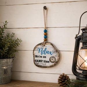 May include: A hanging wooden sign with a distressed white finish, featuring the blue text "Relax" and black text "We're on CAMP time" with a clock graphic. It is suspended by a jute rope adorned with two blue and two brown wooden beads.