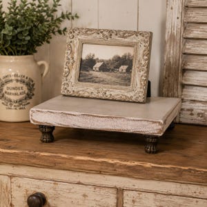 May include: A rectangular wooden riser with a distressed white finish and four dark, carved ball-and-claw feet. Resting on the riser is an ornate, rectangular picture frame with a cream-colored, scrollwork border containing a black and white landscape photo.