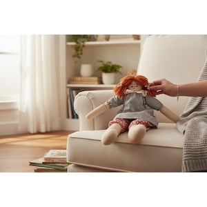 May include: A handmade rag doll with orange yarn hair, a gray checkered dress, and floral print pants. The doll has a white collar and is sitting on a cream-colored armchair. The doll's arms and legs are long and slender.