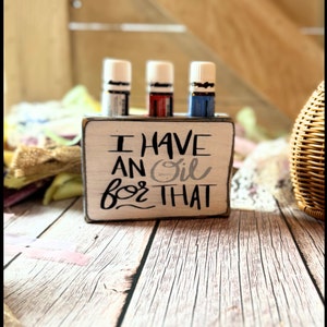 May include: Wooden essential oil holder with the text "I have an oil for that" in black and white lettering. Three essential oil bottles are in the holder. The bottles are white with blue, red, and clear labels.