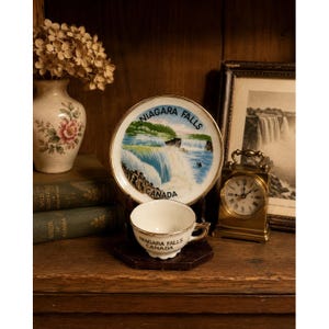 May include: A vintage teacup and saucer set with a Niagara Falls theme. The white porcelain cup and saucer have gold trim and the words "Niagara Falls Canada". A small clock and a vase with dried flowers are also visible.
