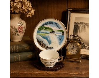 May include: A vintage teacup and saucer set with a Niagara Falls theme. The white porcelain cup and saucer have gold trim and the words "Niagara Falls Canada". A small clock and a vase with dried flowers are also visible.