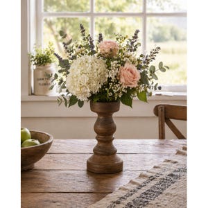 May include: A wooden vase holds a floral arrangement of white hydrangeas, pink roses, and sprigs of lavender and greenery. The vase sits on a wooden table with a patterned runner. A wooden bowl of green apples is in the foreground.