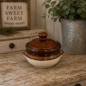 May include: A small, round, two-toned ceramic crock with a lid. The upper portion and lid feature a glossy, dark brown glaze, while the lower portion is a matte, off-white color. The pot includes two small, integrated side handles for gripping.