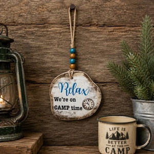 May include: A hanging wooden sign with a distressed white finish featuring the blue text Relax and black text We're on CAMP time with a small clock graphic. The sign is suspended by a jute twine loop adorned with two blue and two brown wooden beads.