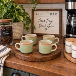 May include: Four light green ceramic mugs with a thin orange rim and horizontal textured stripes, arranged on a round wooden tray. The mugs feature a rounded handle and a smooth finish, providing a consistent look for a coffee station setup.