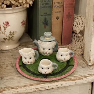 May include: A miniature tea set featuring a teapot, a creamer, and two cups, all sculpted with white cat faces and blue handles. The set sits on a round, green, grass-textured tray with a pink border, arranged as a decorative display piece.