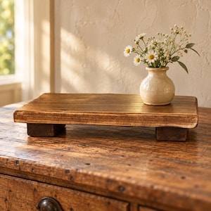 May include: A rectangular wooden riser with a dark brown stained finish, supported by four short block feet. A small, cream-colored ceramic vase containing a bouquet of white daisies and baby's breath sits on the right side of the riser.