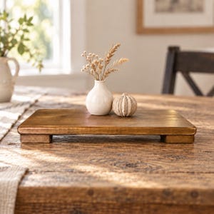 May include: A rectangular wooden riser with a smooth, medium-brown finish and four short feet. Resting on the surface are a small, off-white ceramic vase containing dried beige stems and a round, textured, off-white decorative object.