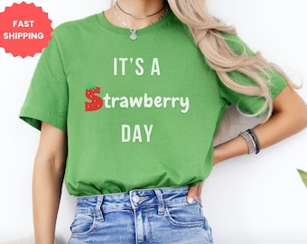 Op de afbeelding: Groen T-shirt met de tekst "It's a strawberry day" in witte letters. De letter "S" is een rode aardbeienafbeelding.