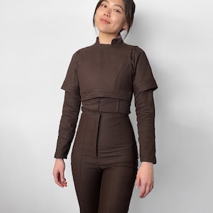 Può includere: Una tuta marrone con scollo alto e maniche lunghe. La tuta ha un corpetto aderente e pantaloni a gamba larga.