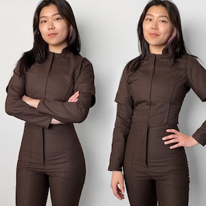 Può includere: Due persone indossano tute marroni a maniche lunghe e con un colletto alto. Le tute hanno una cerniera sul davanti e un passante per la cintura in vita.