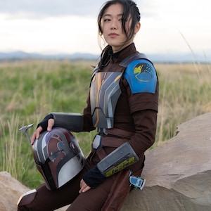 Pu&ograve; includere: Una donna che indossa un costume marrone e blu con un casco in grembo. Il costume ha un design blu e giallo sulla spalla e un design nero e grigio sul petto. Il casco &egrave; nero, bianco e rosso con una visiera blu.