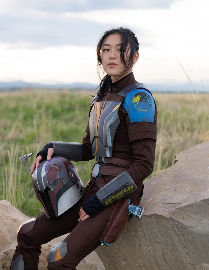 Pu&ograve; includere: Una donna che indossa un costume marrone e blu con un casco in grembo. Il costume ha un design blu e giallo sulla spalla e un design nero e grigio sul petto. Il casco &egrave; nero, bianco e rosso con una visiera blu.