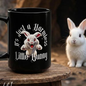 Puede incluir: Taza de cerámica negra con el texto "It's Just a Harmless Little Bunny" y un dibujo de un conejo blanco amenazante. Un conejo blanco real también está presente. La taza está sobre una superficie de madera.