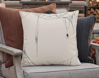May include: A square throw pillow with a cream-colored cover featuring a black line-art illustration of spider webs and a small spider hanging from a thread. The pillow is positioned in front of a textured rust-colored pillow and a dark gray pillow.