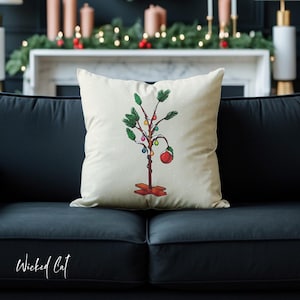 Puede incluir: Un cojín decorativo crema con un diseño caprichoso de árbol de Navidad. El árbol está adornado con luces de colores y un gran adorno rojo. El cojín tiene un patrón de puntos y está colocado en un sofá negro.