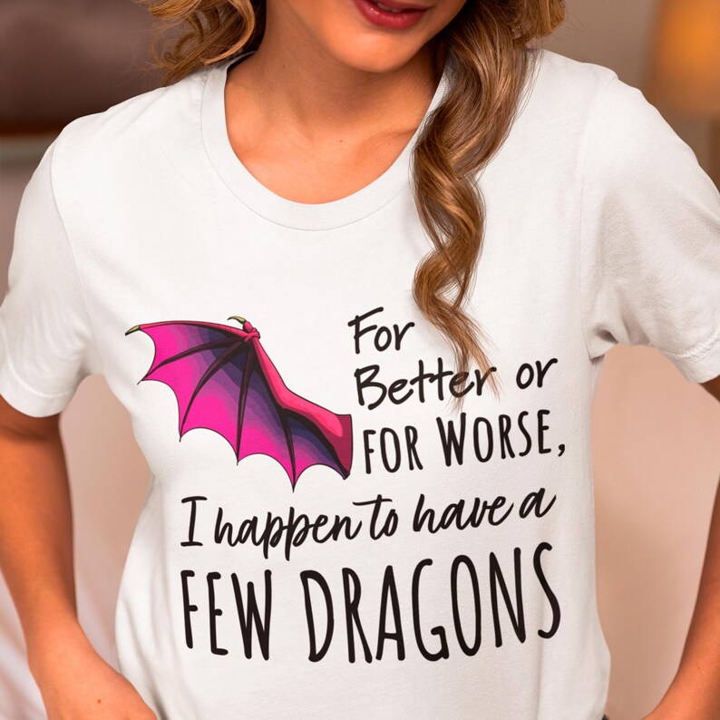 Pu&ograve; includere: Maglietta bianca con un'immagine di ala di drago rosa e viola e il testo "For Better or For Worse, I happen to have a FEW DRAGONS".