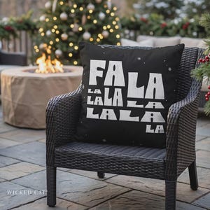 May include: A square black throw pillow featuring the white text FA LA LA LA LA LALA LA with small white snowflake accents. The pillow is positioned on a dark woven outdoor chair, displaying a bold, distressed-style font across the front surface.