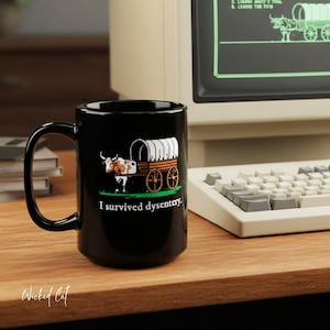 May include: Black ceramic mug featuring a covered wagon graphic with oxen and the text "I survived dysentery." The mug has a large handle and is set on a wooden surface. A vintage computer is in the background.