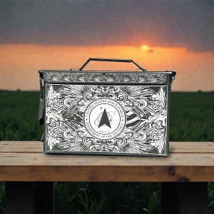 May include: A dark green metal box with a black handle and intricate white designs. The front features the United States Space Force emblem within a decorative frame. The box is set on a wooden surface against a sunset backdrop.