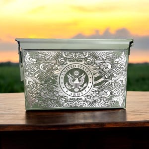 May include: A green metal ammo can with a silver embossed design featuring the United States Army seal and wings. The can has a hinged lid and a carrying handle.