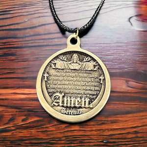 May include: A gold-toned pendant necklace with a round charm featuring a prayer inscribed in English. The charm has a raised image of Jesus with outstretched arms and the words "Amen" and "Serenity" inscribed below.