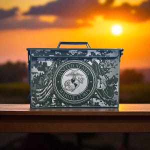 May include: A rectangular metal box with a camouflage pattern and a black handle. The front of the box features the United States Marine Corps emblem, with the words "United States Marine Corps" encircling the emblem and two flags.
