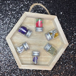 Peut inclure: Un plateau hexagonal en bois avec six petits pots en verre contenant divers objets, dont des fleurs séchées, des paillettes et du papier. Les pots ont des couvercles argentés et sont disposés en cercle.