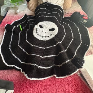 Puede incluir: Una manta de ganchillo en blanco y negro hecha a mano con la cara de Jack Skellington de Pesadilla antes de Navidad. La manta tiene un dise&ntilde;o circular con rayas blancas y una cara blanca. Se ve un ovillo de hilo negro.