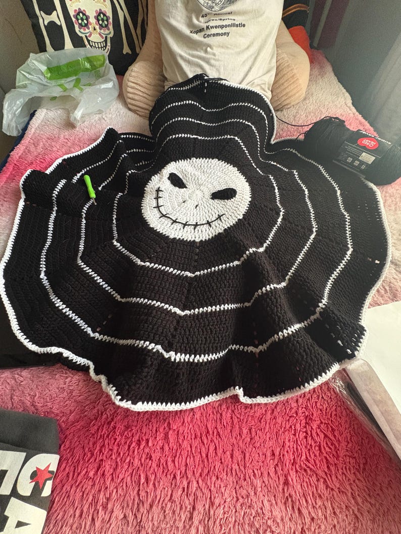 Puede incluir: Una manta de ganchillo en blanco y negro hecha a mano con la cara de Jack Skellington de Pesadilla antes de Navidad. La manta tiene un dise&ntilde;o circular con rayas blancas y una cara blanca. Se ve un ovillo de hilo negro.