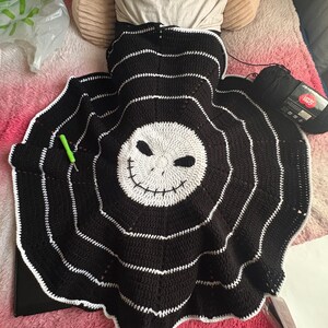 Puede incluir: Una manta de ganchillo en blanco y negro hecha a mano con una cara de calavera sonriente en el centro. La manta tiene c&iacute;rculos conc&eacute;ntricos con costuras blancas y un borde festoneado. Se ve un ganchillo verde y un ovillo de lana negra.