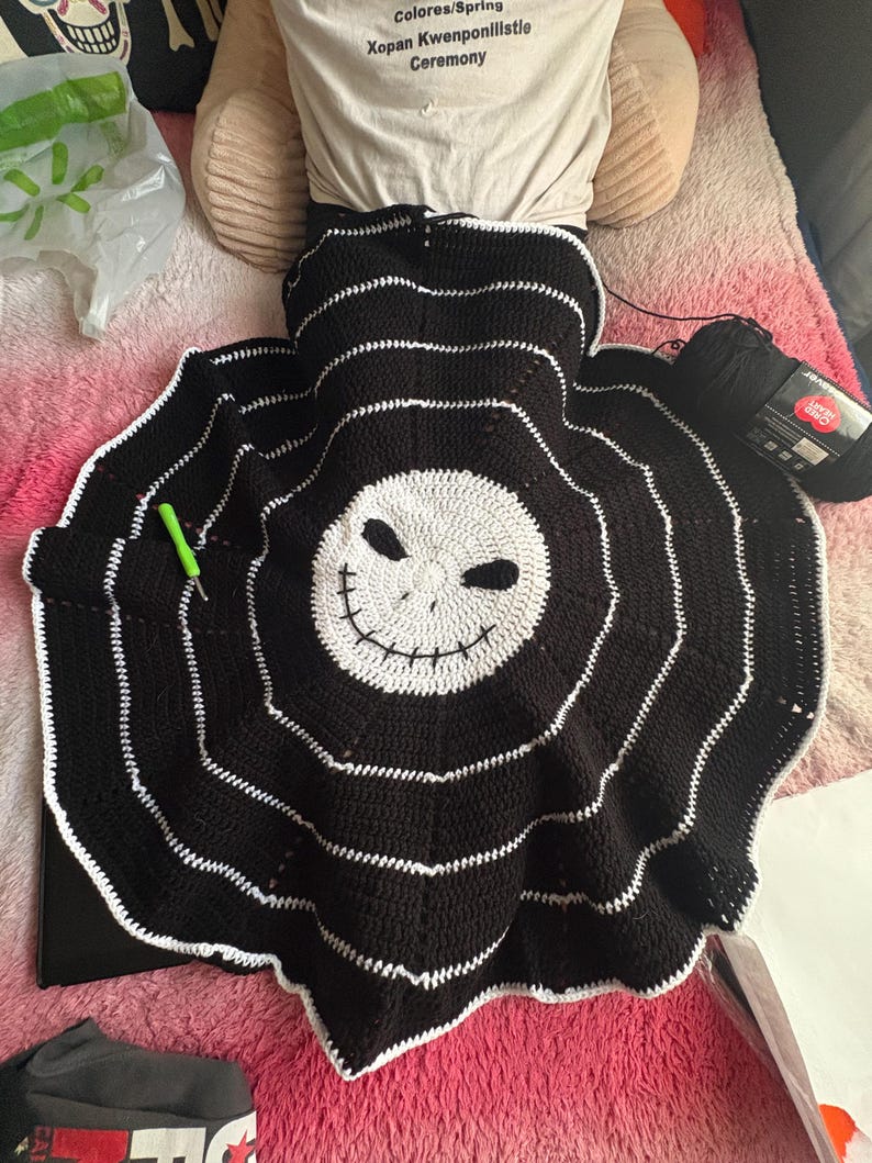 Puede incluir: Una manta de ganchillo en blanco y negro hecha a mano con una cara de calavera sonriente en el centro. La manta tiene c&iacute;rculos conc&eacute;ntricos con costuras blancas y un borde festoneado. Se ve un ganchillo verde y un ovillo de lana negra.