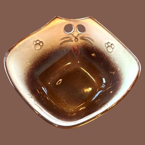 May include: A brown and white ceramic cat-shaped bowl with a cat face and paw prints. The bowl has a handle on the top.