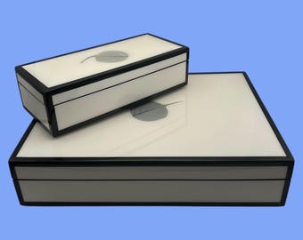 May include: Two rectangular white boxes with black trim. The top of each box has a leaf design. The boxes are stacked on top of each other. The background is a solid light blue.