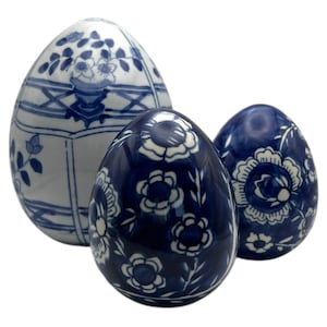 May include: Three ceramic decorative eggs in varying sizes. The eggs are primarily blue and white, featuring floral and geometric patterns. The largest egg has a detailed floral design and a vase motif. The smaller eggs have a floral pattern.