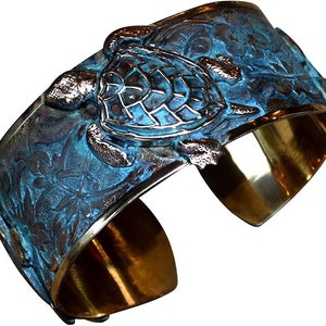 May include: A wide cuff bracelet featuring a raised sea turtle motif on the outer surface. The exterior has a textured, oxidized blue-green patina finish with intricate floral patterns, while the interior is a smooth, polished metallic gold tone.