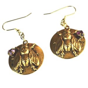 May include: A pair of gold-colored bee earrings. Each earring features a detailed bee design on a circular pendant, with a small purple gemstone accent. The earrings have a hook closure.
