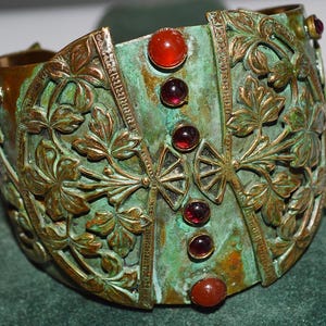 May include: An antique cuff bracelet with a verdigris finish. The bronze-toned metal is adorned with floral and leaf patterns. The bracelet is embellished with red and maroon gemstones.
