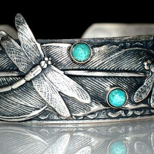 May include: A silver cuff bracelet featuring an embossed feather pattern, two detailed dragonfly motifs, and two small, round, turquoise-colored stones set into the metal. The silver has an oxidized finish to highlight the intricate textures.