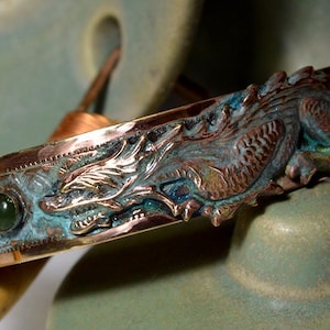 May include: A copper cuff bracelet featuring a raised dragon relief with a weathered teal patina finish. The band is accented with two smooth, round, dark green cabochon stones set into the metal, showcasing a textured and metallic appearance.