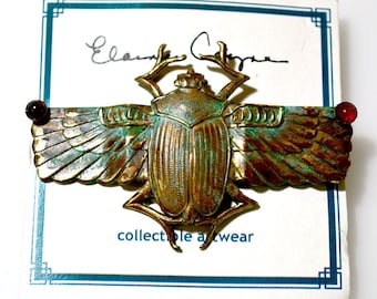 May include: A bronze scarab beetle with wings and red glass accents. The beetle is on a white card with the text "Elaine Coyne" and "collectible art wear".