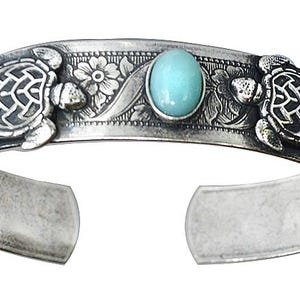 May include: A silver cuff bracelet featuring two embossed turtle figures, floral engravings, a central oval light blue stone, and two small green circular stones on the ends. The metal surface has a darkened, oxidized finish in the recessed areas.