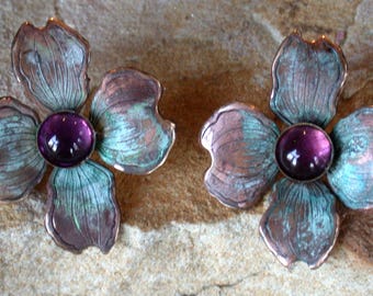 May include: A pair of flower-shaped earrings featuring four petals with a weathered copper finish in shades of teal and brown. Each earring has a smooth, round, deep purple cabochon stone set in the center, mimicking the appearance of a blossom.