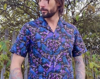 Pode incluir: Camisa de manga curta com fundo escuro e padrão de pássaros azuis e detalhes florais roxos. A camisa tem gola clássica e é feita de tecido leve.