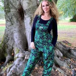 May include: Overalls with a green and black pattern, worn with a black long-sleeved top and burgundy boots. The overalls have a unique design. A leafy headband is worn. The background features a large tree in a natural setting.