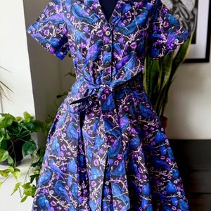 May include: A short-sleeved dress with a black background, featuring a pattern of blue birds and purple flowers. The dress has a collared neckline, a button-down front, and a matching tie belt. The skirt is pleated.